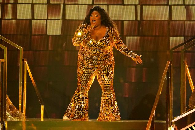 A photo of Lizzo in a shimmering gold outfit on a stage performing in summer 2022