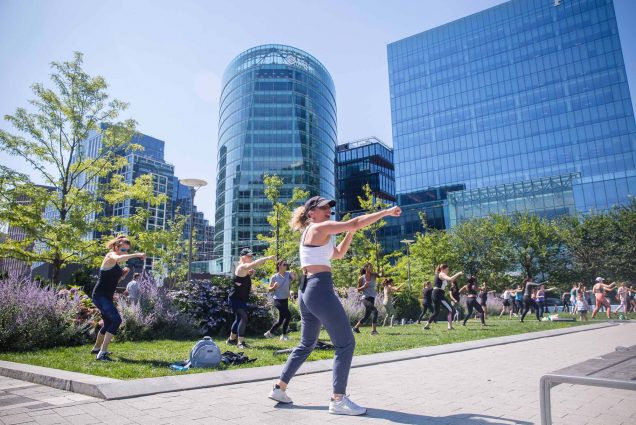 Photo of a women wearing an athletic crop top and leggings punching the air as she speaks into a wireless headset on a bright, sunny day. A group of people in athletic wear can be seen copying her punch as they stand on the green lawn behind her. City buildings can be seen looming in the distance.