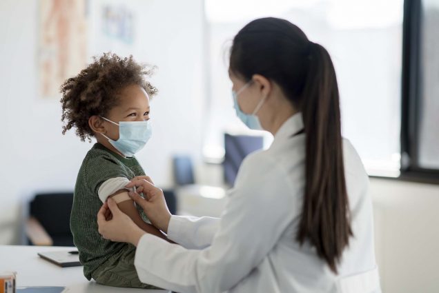 Photo of a Black toddler with brown curly hair smiling from underneath their mask as a doctor puts a bandaid on their arm after giving them a vaccine. The baby wears a green shirt and sits on a doctor's table. The doctor is a youngish AAPI woman will a long, brown ponytail wearing a white doctor's coat and face mask. Doctor's office equipment is seen blurred behind them.