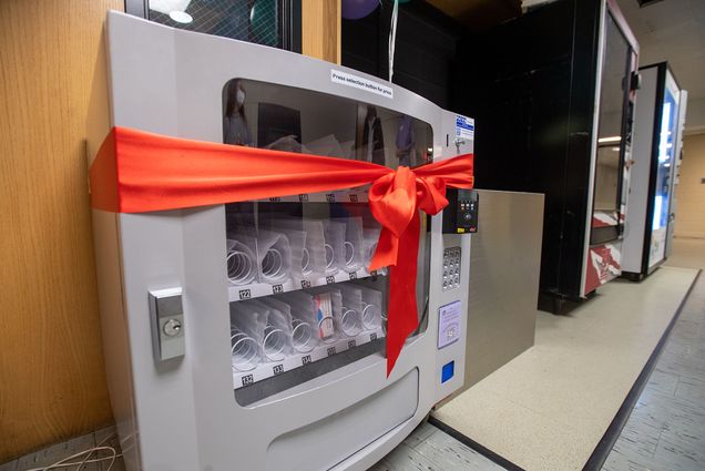 Photo of a small grey vending machine. Most of the vending slots are empty in the photo, except for a few that have plan b in them. A red ribbon is tied around the machine.