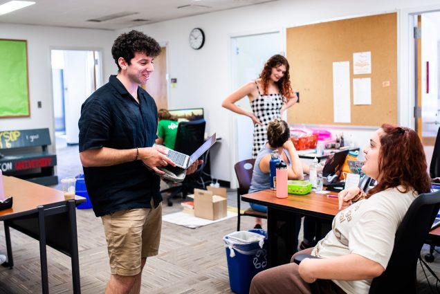 Photo of Jacob Nesson (left) talking with Sam Perez (CAS 25) about the First Year Student Outreach Project. A young man in a black tee and khaki shorts talks to a young woman sitting in a chair in front of him. Two other young woman can be seen chatting behind them. Theyre all in a small office space.