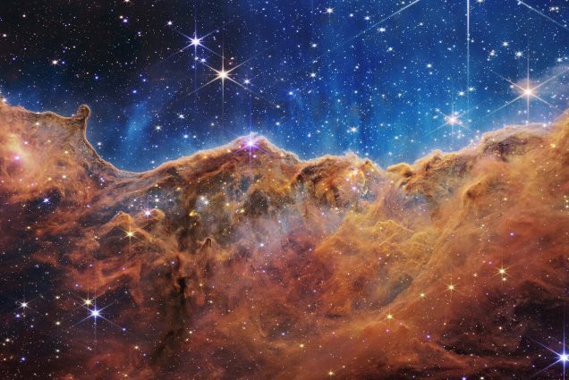 Image: Photo of the Carina Nebula. Image looks like craggy mountains on a moonlit evening with an ombre black to blue "sky" filled with stars and a cloud of orange-y red haze/cloud.