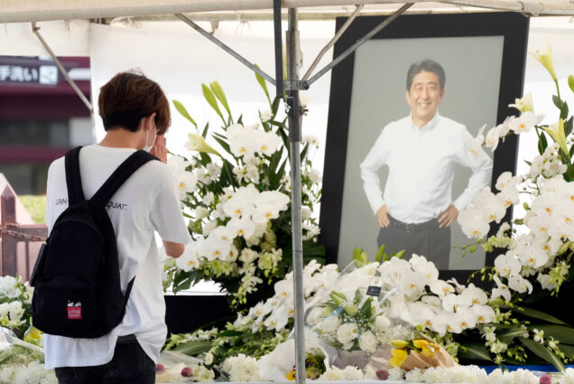 photo of a young Japanese person offering flowers and bowing their head in prayer in front of a black-framed photo of former Prime Minister Shinzo Abe, at Zojoji temple prior to his funeral wake Monday, July 11, 2022, in Tokyo. In the photo, Abe wears a white button down shirt, black pants, and smiles with his hands on his hips. Large, white flower displays are seen around the photo and on the table in front. The saddened young person wears a white shirt and a black backpack and has short light brown hair.