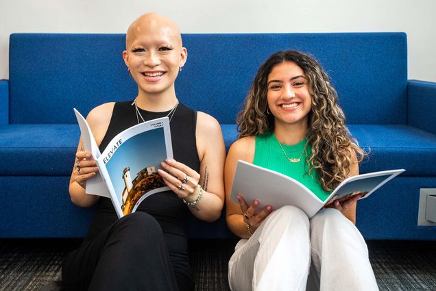 Photo of Elevate editors-in-chief Kat Quach (COM’24) (left) and Valerie Sanchez Covaleda (CGS’22, COM’24) with copies of their magazine in the Newbury Center. Both sit on the floor in the newbury center, leaning against a blue couch and holding first issues of Elevate open in their laps. Quach is a young Vietnamese-American and trans woman who wears a black blouse and small earrings; Quach has a shaved head and has long black lashes. Sanchez is a young, women of color with long brown hair with blonde highlights. She wears white pants, a green blouse, and gold necklack. Both smile.