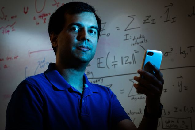 Photo of Mayank Varia, associate professor in the Faculty of Computing & Data Sciences, posing for a photo in front of a whiteboard filled with various equations. A middle-aged man stands in dark room and holds a white cell phone up. He, the phone, and the whiteboard background are lit up by light blue neon light.