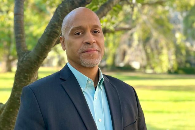 Photo of Scott Taylor. A bald black man with a neatly trimmed silver beard and mustache poses and looks directly at the camera. He wears a navy blazer and teal collared shirt and stands in front of a tree in a green field.