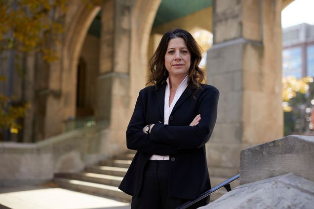 Photo of Jessica Silbey standing in a courtyard with stone arches. A middle-aged woman with shoulder length brown hair wears a black suit ensemble and stands with arms crossed.