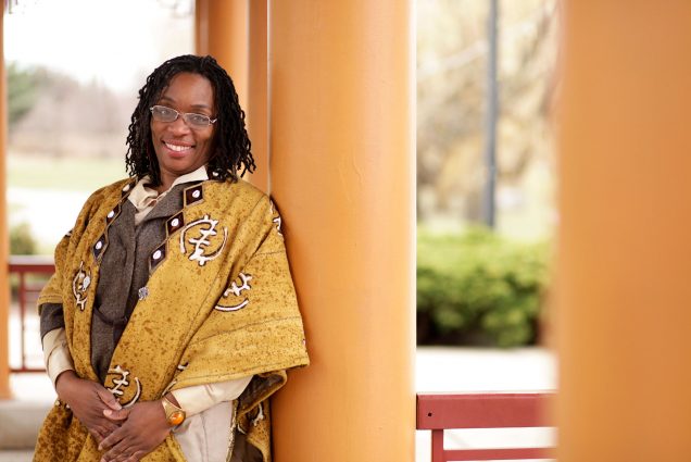 Photo of Deidra Somerville. A black woman with dreadlocks and glasses leans against a burnt yellow pillar and smiles for the camera. She wears a brown collared shirt and a yellow shawl with a Ghanaian adinkra pattern.