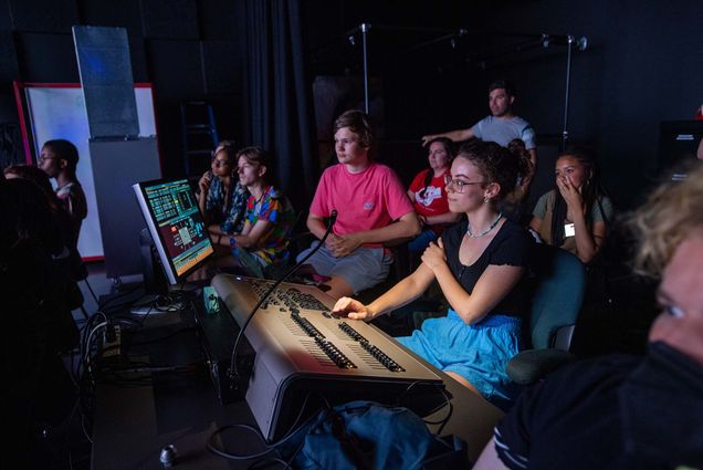 Photo of Nora Kempner (center) a 17-year-old high school student from Medway, Mass., who is enrolled in the BU Summer Theatre Institute’s design program. Kempner is a young White woman with wavy hair, pulled back, wearing a black t-shirt and blue shorts. She sits at a table with lighting controls and a screen and smiles as she shows her lighting scene design with classmates. Other students can be seen seated behind her in the darkened room.