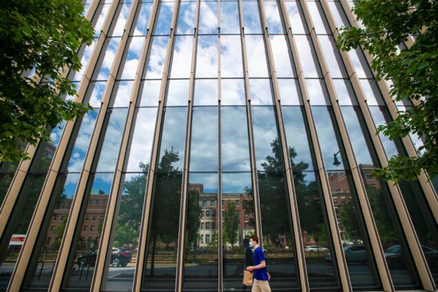 Photo of Cloudy blue skies reflecting in the windows at the BU Kilachand Center. A long figure walks by on the sidewalk.