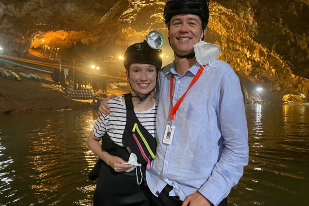 Photo of Michael Russell Gunn (COM’07) and Tristan Roche (COM’20) visiting the set of Thai Cave Rescue in the Tham Luang Nang Non cave in northern Thailand. The two stand in a cave giving one armed hugs as they wear helmets with head flashlights on them and smile for the camera.