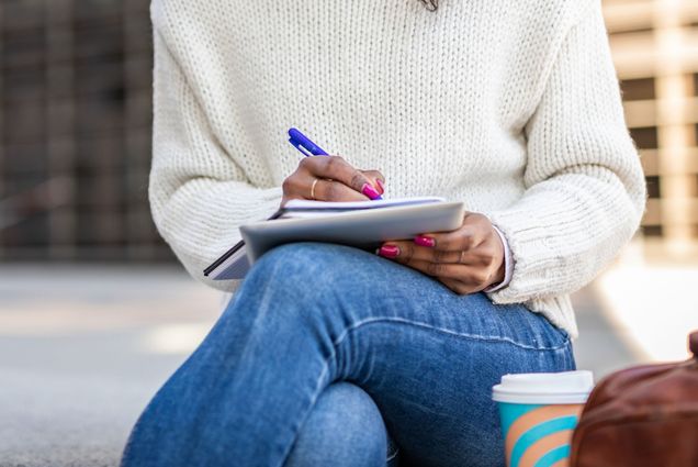 a person wearing jeans and a sweater with their legs crossed writes in a journal outside on a sunny day