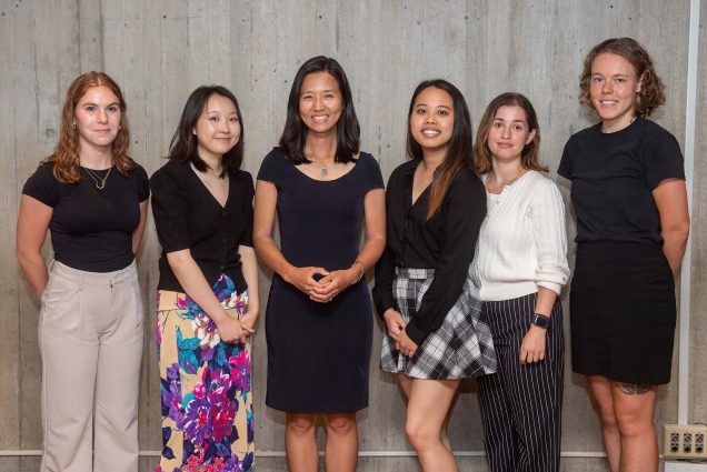 Photo of Boston Mayor Michelle Wu (third from left) with five of this year’s BU City Scholars Summer Fellows: Willa Nessoff (CAS'24) (from left), Nancy Wang (Questrom'24), Connie Situ (CAS'23), Shirli Marini (Questrom'24), and Stella Dzialas (CAS'24). Wu is a middle-aged AAPI woman with shoulder length black hair who wears a black dress. Nessoff, a White woman, wears a black shirt and flowy tan pants, has wavy reddish hair. Wang is an AAPI woman with shoulder-length black hair who wears a black v-neck and a floral skirt. Situ is a woman of color with long brown and blonde ombre hair who wears a black long sleeve shirt and a black and white plaid shirt. Marini is a light-skinned woman with short strawberry blonde hair who wears a white sweater and black and white speckled pants. Dzialas, a White woman, wears a knee-length black dress and has short, wavy dirty blonde hair. They pose in front of a cement wall.