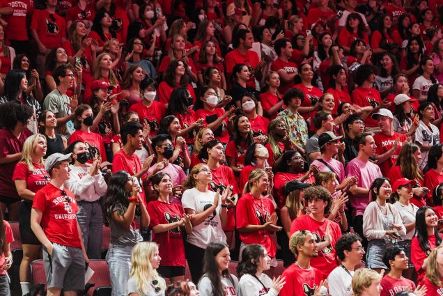 Photo of a diverse group of students clapping during the matriculation event in Agganis Arena. Almost all of them wear red BU tshirts, with either the words "Boston University" or an image of a terrier on them. Many also hold papers in their hands.