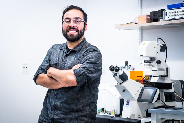 Photo of Steve Ramirez, a CAS assistant professor of psychological and brain sciences, smiling and posing with arms crossed in a white lab. He wears black glasses. and a dark grey collared shirt.