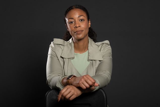 Studio portrait of Amanda Bailey. A black woman with straight black hair and a light smile leans on a chair with hands intertwined on top of a black chair. She wears a black and white blouse and no-frame glasses.