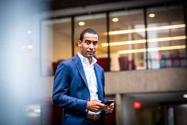 Photo of Trevor Rozier-Byrd (LAW'10) posing for a photo. A dark, tan man wearing a blue blazer and pants and white collared shirt holds his black cell phone in hand as he looks to the camera.