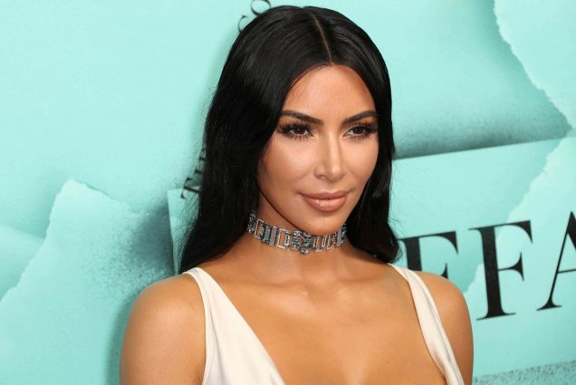 Photo of Kim Kardashian posing in front of a light blue photo area. A tan woman with long black hair and wearing a thin metal choker and white top, smiles slightly for cameras.