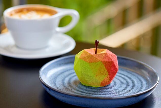 Photo of a delicate, geometric apple- shaped dessert. Apple is a light green and red. A blurred cup of coffee is also shown on the table in the background.
