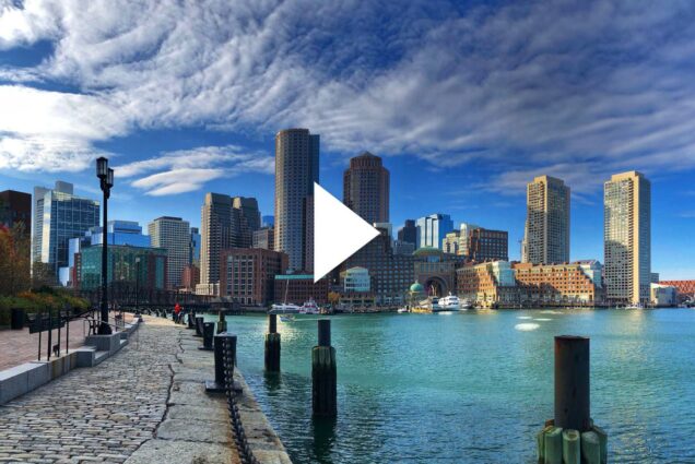 Photo of Boston's Seaport district. A stone walkway on the left lines a large body of water as the Boston seaport cityscape is scene in the background. A white triangle play button is overlaid in the center.