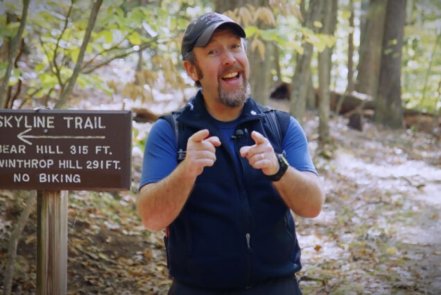 Photo: Hutch Hutcherson stands and points to the camera with both hands. He wears a blue short sleeved shirt, and black puffer vest and cap as he stands on a hiking trail. Wooden sign behind him gives details on the trail he is on.