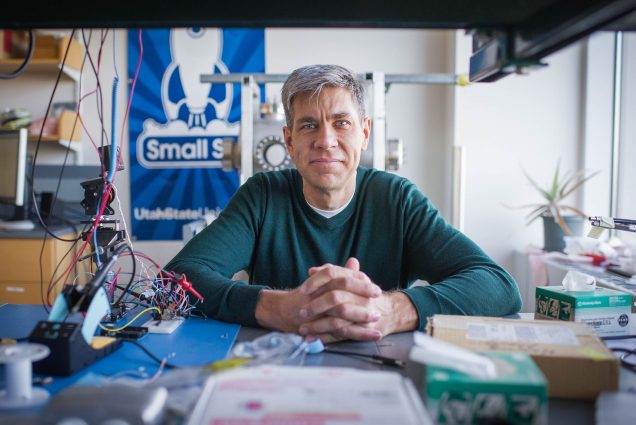 Photo: ENG professor of electrical and computer engineering, Josh Semeter poses for photo in his lab on Wednesday, January 20, 2016. A white man with silver grey hair sits with hands folded in front of him and resting on a desk cluttered with papers, wires, and various hardware constructs. He wears a dark green sweater. A lab filled with random mechanics is shown behind him as a large blue and white spaceship banner hangs on the wall behind him.