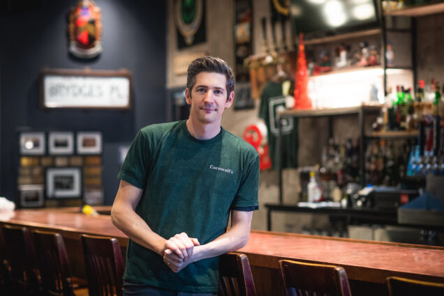 Photo: Cornwall's bartender Bill Moran poses for a photo at the British pub, Cornwall’’s on November 30, 2022. A tall, white man wearing a dark green shirt that says "Cornwall's" on the breast pocket, leans against a wooden bar stool at the counter of a bar.