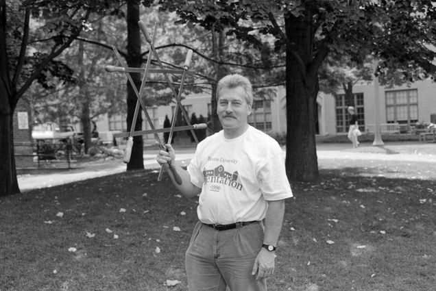 Black and white photo of Anthony Barrand. A white man wearing a white tee shirt that reads "Boston University Orientation 1996" tucked into slacks holds up in one hand a large, wooden jewish star. He stands on a BU lawn on campus.