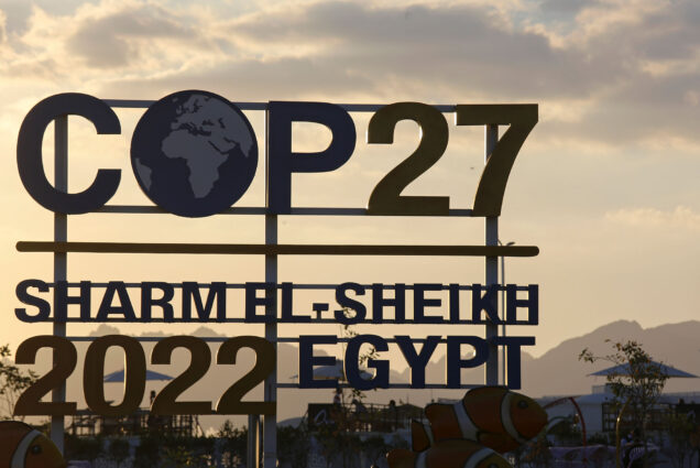 Photo: The sun sets behind signage for the COP27 U.N. Climate Summit in Sharm el-Sheikh, Egypt, Wednesday, Nov. 9, 2022. The large sign reads "COP27, Sharm el-Sheikh, 2022 Egypt" and is shown in shadow against the setting sun. A few large clown fish figures can be seen below the sign.