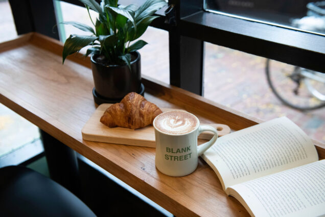 Photo: A large latte sits in a "Blank Street" mug on a wooden counter near window. A croissant sits on a wooden board next to it and an open book lies on its other side.