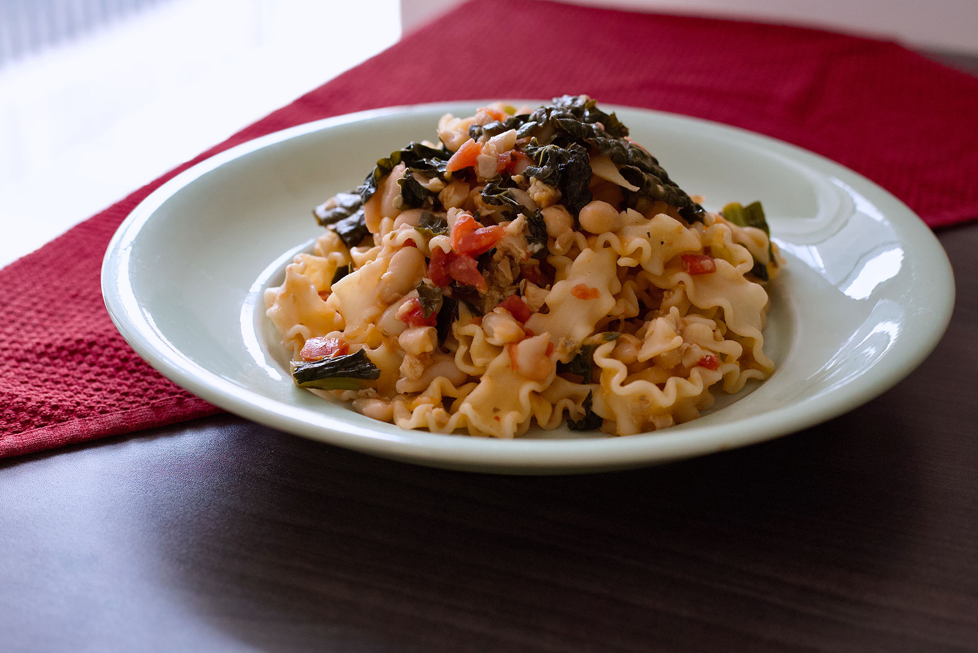 Photo: A pasta dish made of clams, white Beans, Tomatoes, and kale in a red sauce is shown plated on wooden table and red cloth.