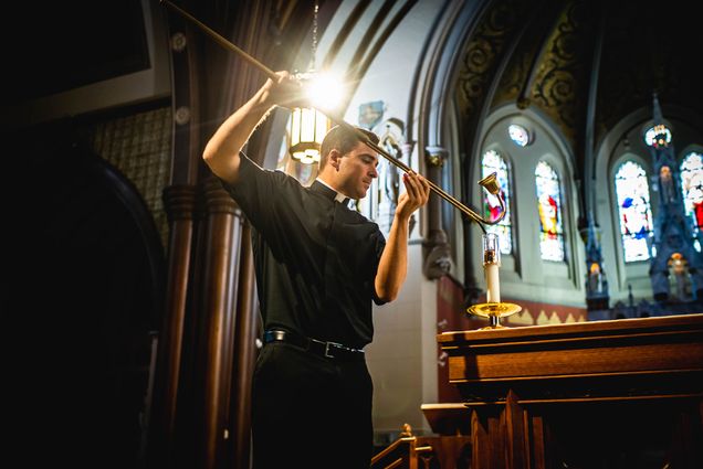 Photo of LeBlanc dressed in black priest outfit lighting a candle. Stained glass windows are seen behind him.