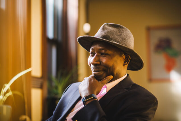 Photo: Fallou Ngom, professor of anthropology, poses for a photo on November 22, 2022. A black man wearing a grey medium-brimmed hat, light pink collared shirt, and navy blue blazer, poses with hand on chin and a soft smile as he sits in a chair.
