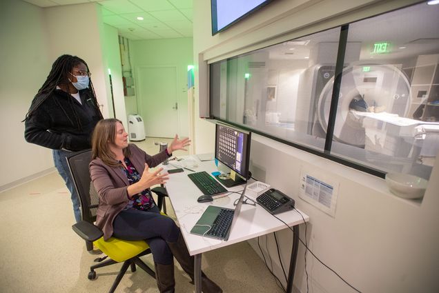 Photo: Associate director of the Cognitive Neuroimaging Center Stephanie McMains (GRS’06), seated, with Arielle Moore (GMS’26) in the Cognitive Neuroimaging Center in CILSE Dec 1. A white woman sits in a chair gesticulating as she sits in front of a table in front of window that shows the fMRI room. A black woman wearing a blue face mask stands behind her listening attentively.