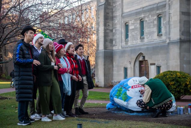 Sam Weinberger (CFA’21) paints the BU rock with holiday flair as the student a cappella group the Allegrettos watches and sings on. A group of students wearing holiday hats and scarves sing as a young man bends down to spray paint a giant rock with a snowman holiday scene.