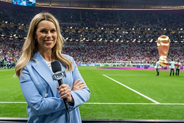 Photo: Fox Sports reporter Jenny Taft on the sidelines at the November 25 FIFA World Cup USA-England match. A white woman wearing a a white shirt and light blue blazer holds up a microphone with a large "FOX" label on it. A green football pitch. The seats are filled with fans in the background and a large replica 2022 World cup can be seen in the background as well.