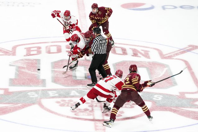 Photo: BU Men's ice hockey faces-off against BC. Various BU hockey players grapple and push on the ice against BC players to get the puck. A referee stands in the center of the group.