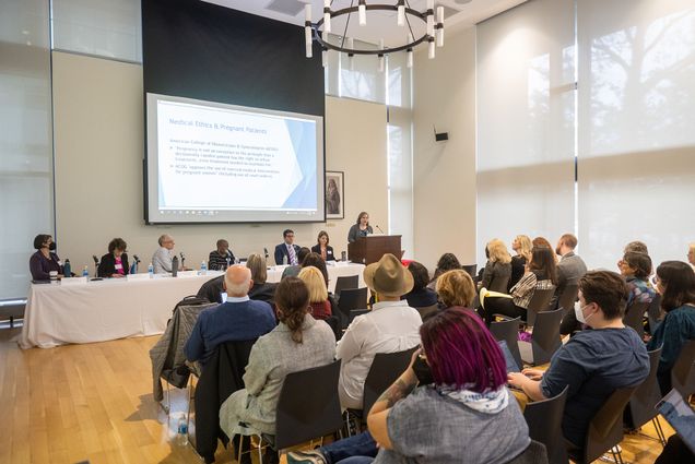 Photo: Photo of the "After Roe and Dobbs: Seeking Reproductive Justice in the Next Fifty Years" event. A panel of speakers are seated at a long white table at the front of a large room. A speaker stands at a podium to the right of the table and a large projector screen hangs down behind them all, showing the screen with a slide that is title "Medical Ethics & Pregnant Patients". A small crowd of people are seated in front of the panel and speaker as the audience.