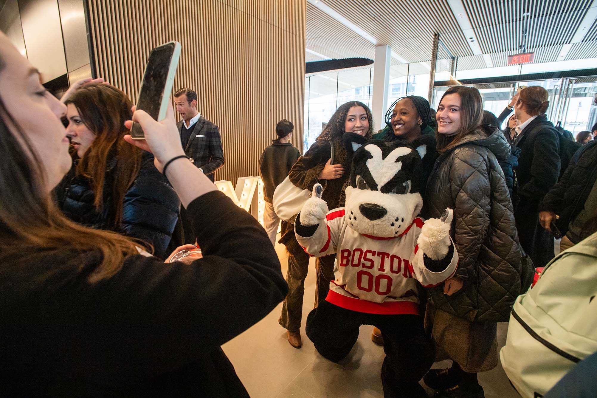 Photo: Students pose with Rhett in the lobby of the Center for Computing & Data Sciences on the first day of classes January 19. Three students stand behind a Boston Terrier mascot wearing a BU Hockey jersey as a woman snaps a photo of them on her phone.