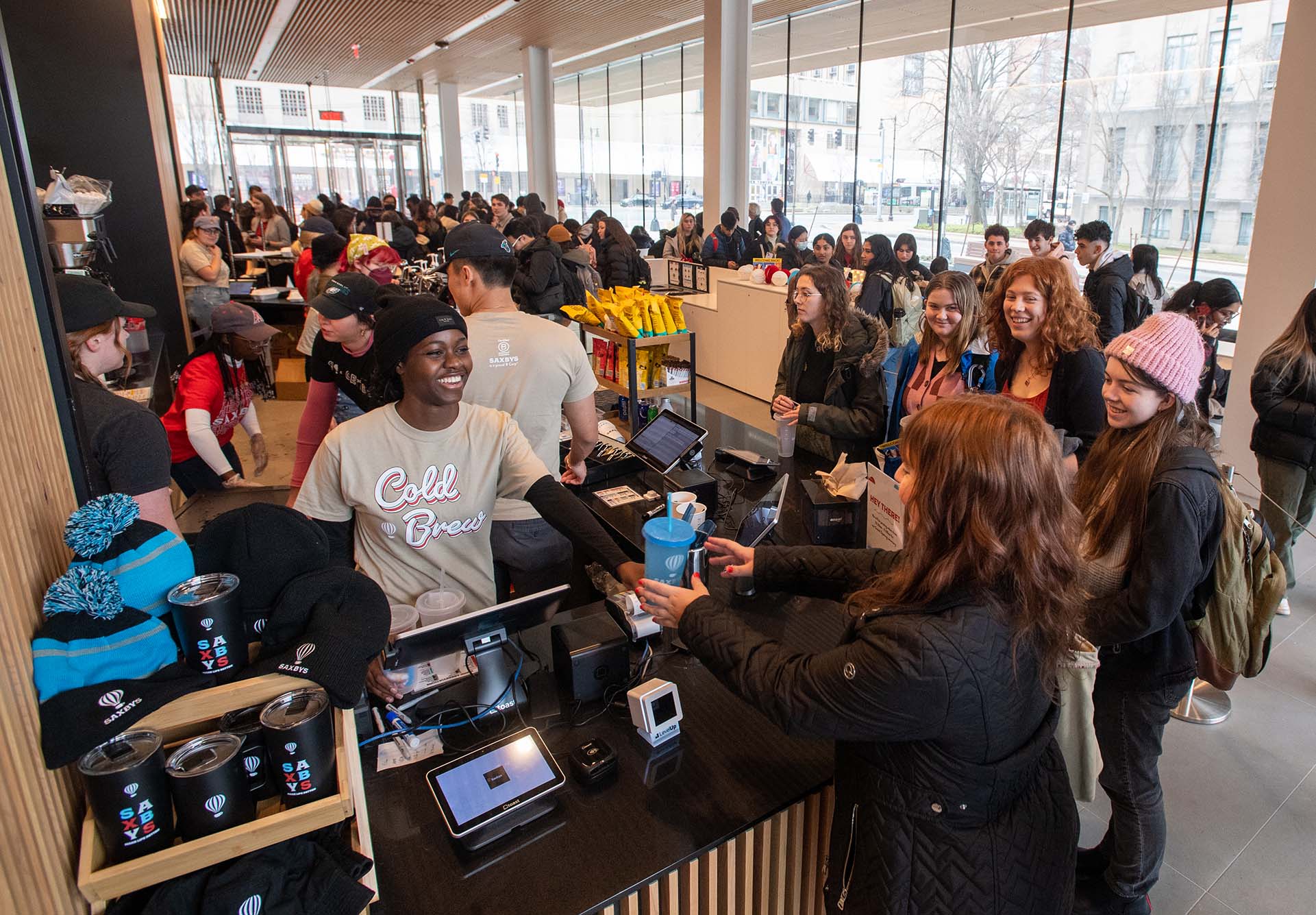 Photo: Malaika Uriesi (CGS’25) at left, hands out free cups at Saxby’s in the Center for Computing & Data Sciences on opening day January 19. A young Black woman wearing a gray Saxby's shirt hands out a drink behind a coffee bar counter. A large crowd is seen around the counter.
