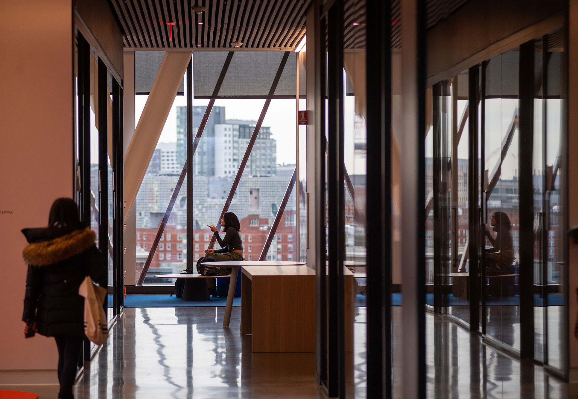 Photo: A student is shown sitting and speaking on her cellphone in front of a large window wall. Image shows off the architecture and design of the interior of the CCDs building.