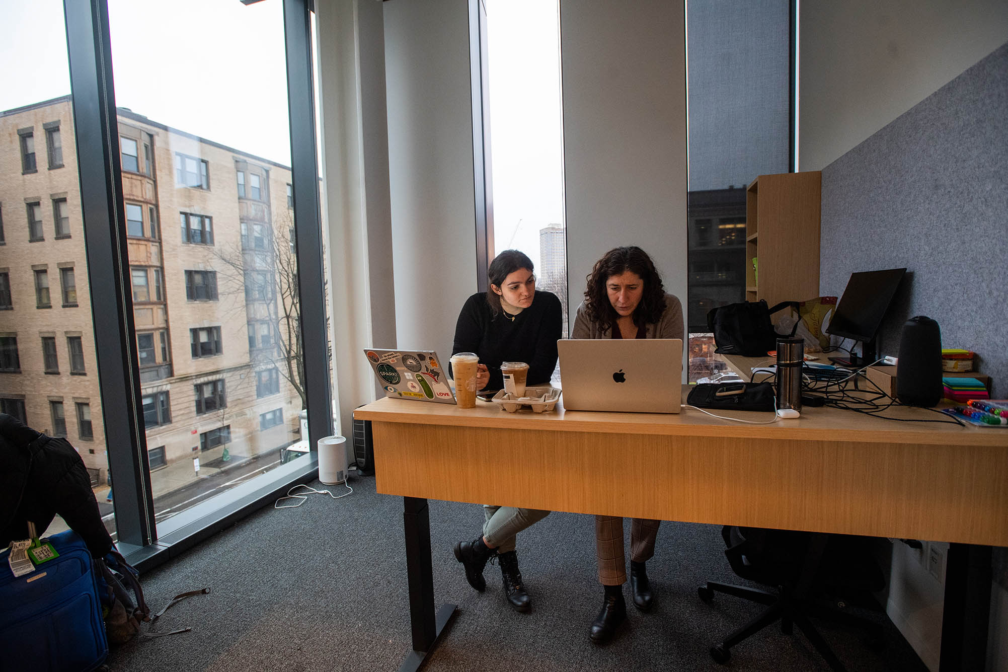Photo: Director of BU SPARK Ziba Cramer, right, and associate program manager Savannah Majarwitz (CAS’22) attend a zoom meeting in Cramer’s new office on the second floor. Two woman sit side by side at a long wooden desk with laptops open. A large window to their left lets in a lot of sunlight.