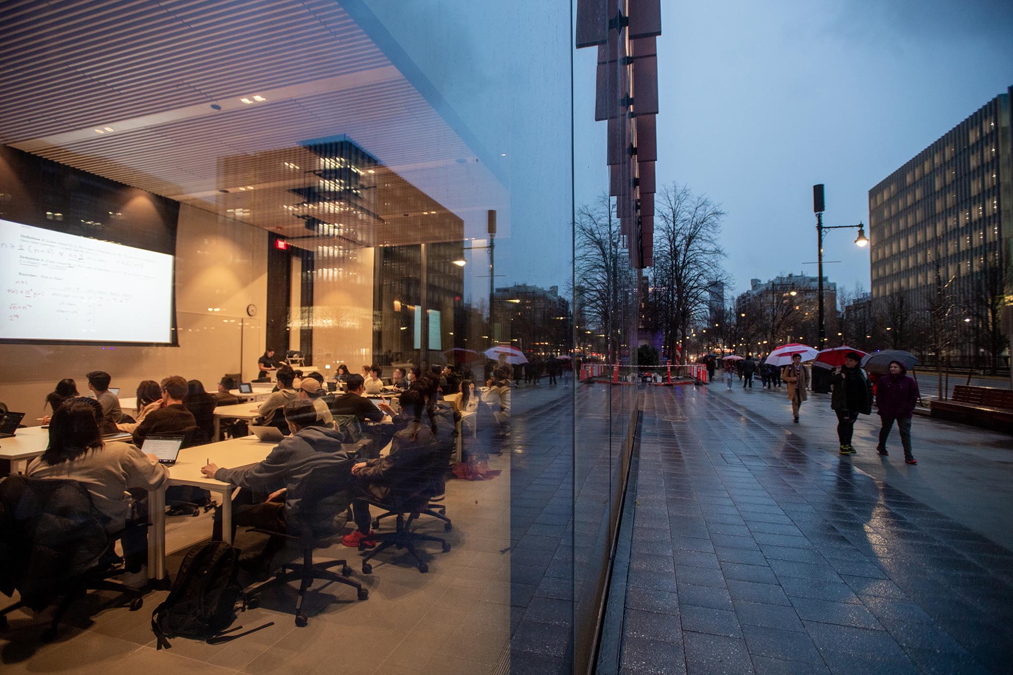 Photo: A side shot of the Center for Computing and Data Science building. On the left you can see a class of students in the midst of a lecture through the glass wall. On the right you see pedestrians walking down the sidewalk with umbrellas during the rainy evening.