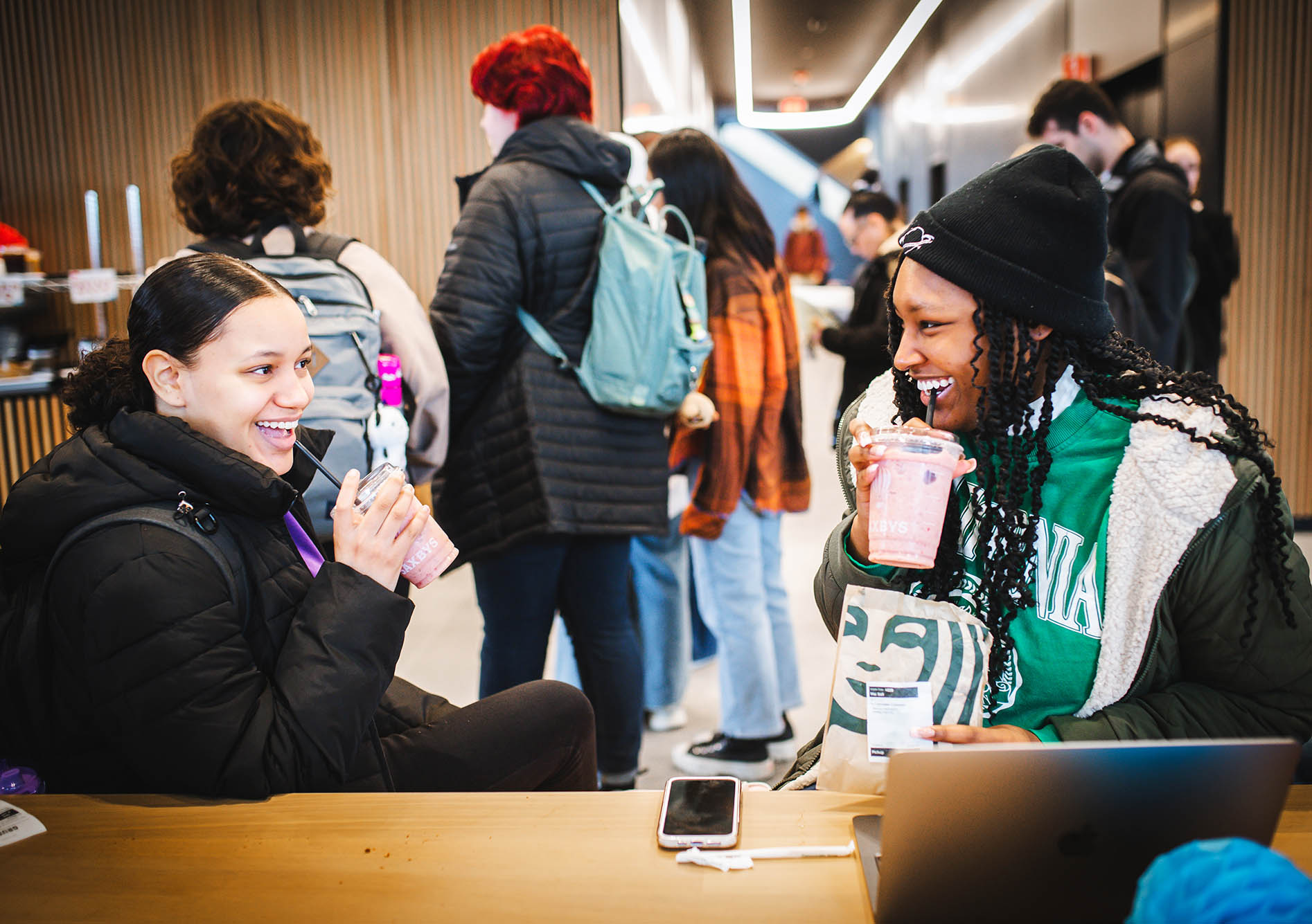 Photo: Dani Duran (SAR 25) and Maimouna Sall (SAR 25) A young light-skinned woman (left) and a young Black woman with long twists (right) smile as they enjoy pink smoothies in front a crowd of students.