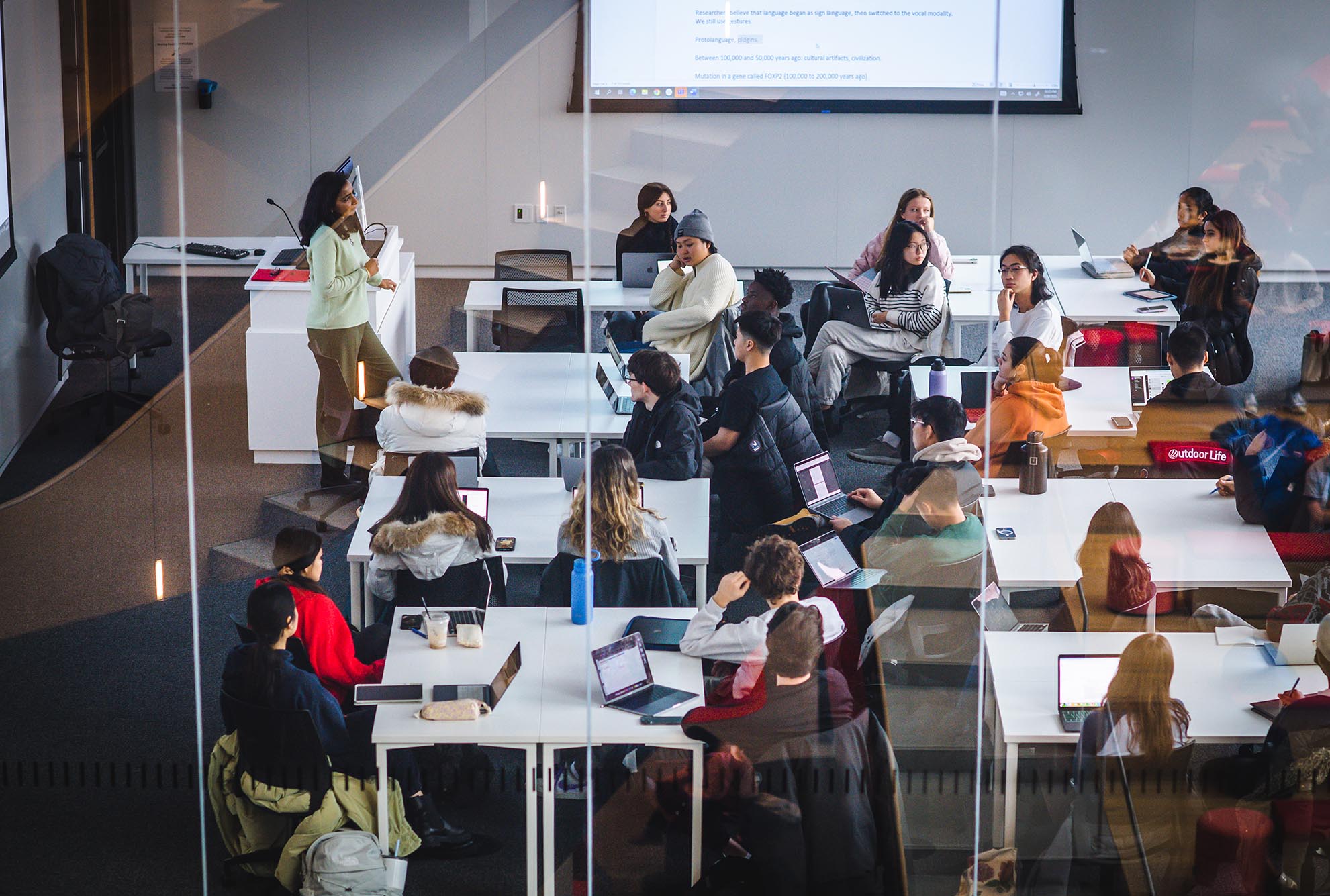 Photo: A professor stands and lectures at the front of a classroom as student seated in groups at square tables look and listen on. Photo is taken form other side of a glass wall.