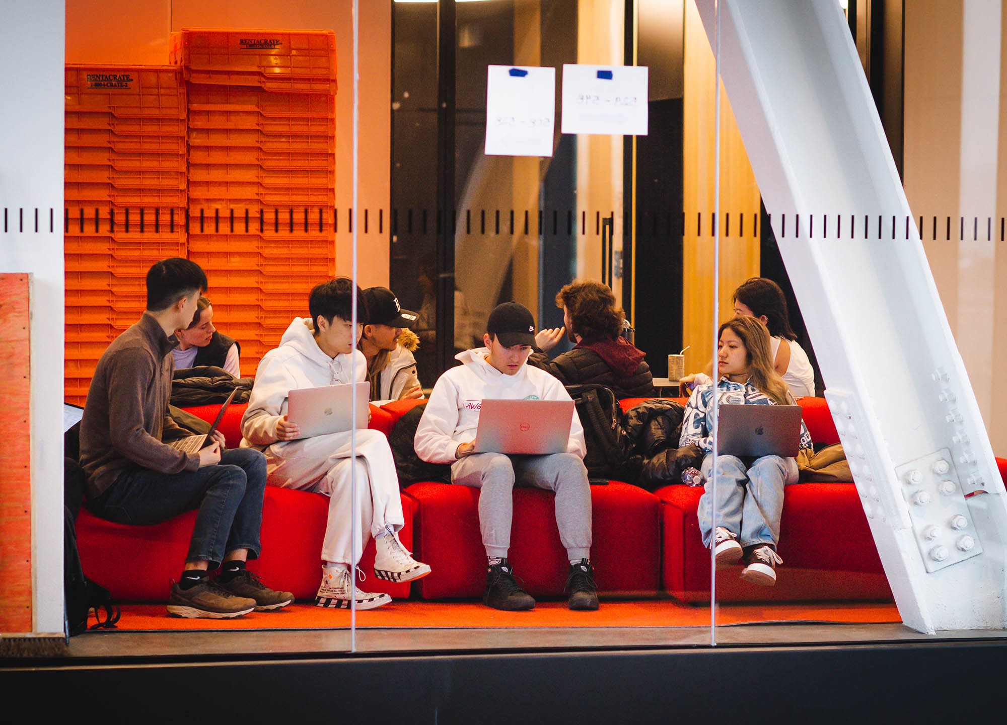 Photo: Students gather in the open spaces of CCDS. A group of students are seen sitting on a large red modular couches through a glass wall.