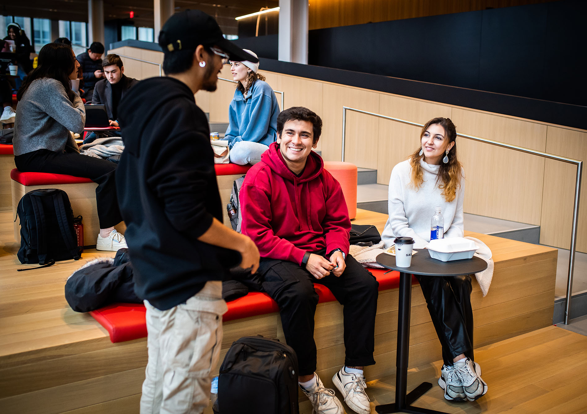 Photo: (Left to right) Perry Sullivan (COM 24) Jonathan Suarez (CAS 25) and Asya Dente (CAS 25) chat in the common during the first week of classes at CCDS on January 20, 2023. Three students sit and chat at the bottom of a large set of wooden, collaborative stairs.