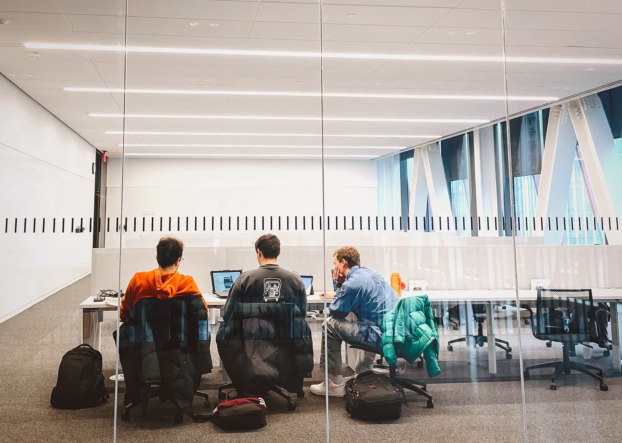 Photo: Students are seen at the other side of a glass wall working together on a project. Three students sitting in chairs with laptops open on desks in front of them look together at one screen in the middle.