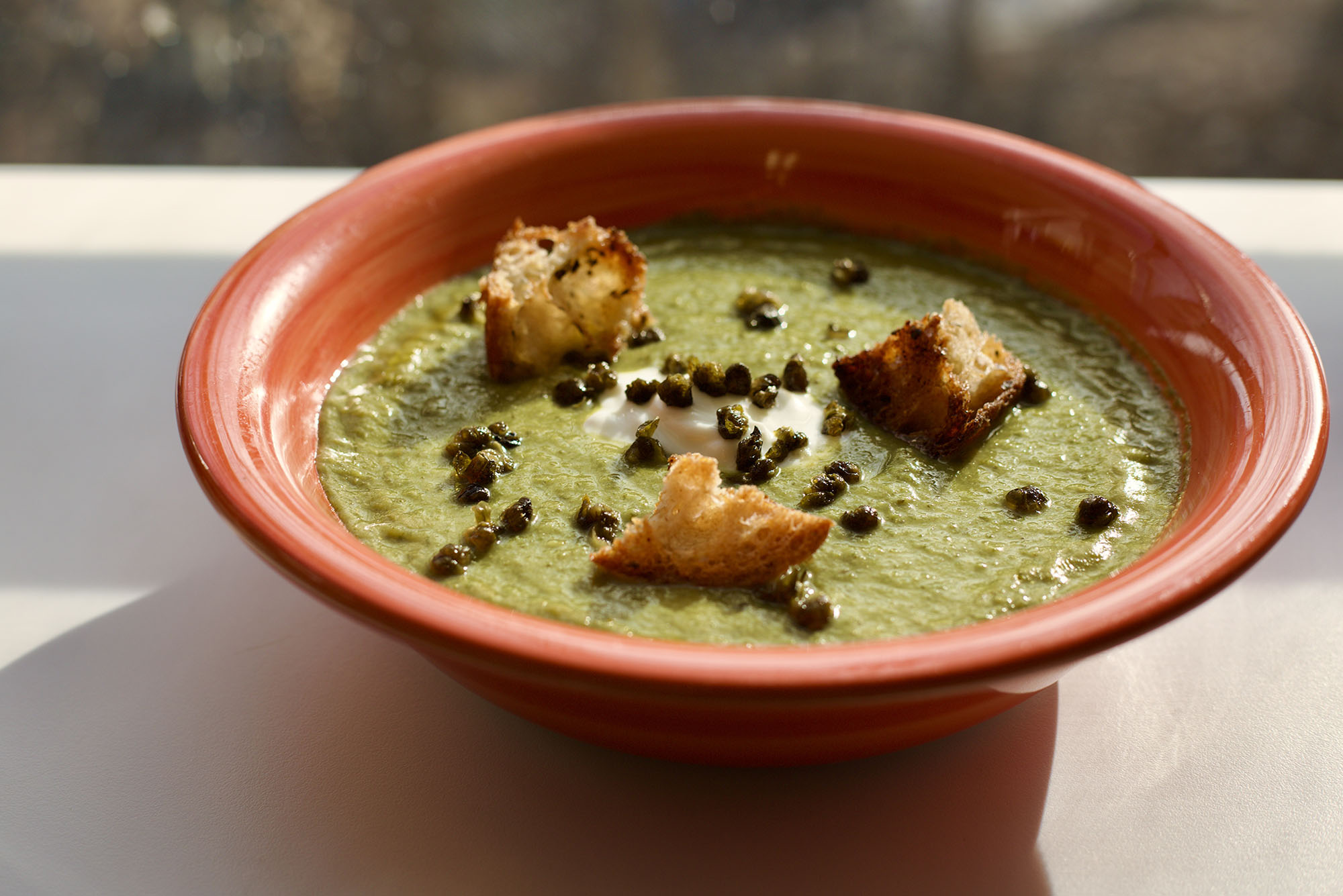 Photo: a red bowl sat next to a window letting in natural light is filled with a dark green soup and topped with homemade croutons, a dollop of cheese, and capers.
