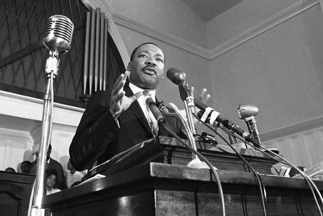 Photo: In this 1960 file photo, Rev. Martin Luther King Jr. speaks in Atlanta. Black and white photo shows a Black man wearing a suit speaking at a podium with hands raised as he speaks.